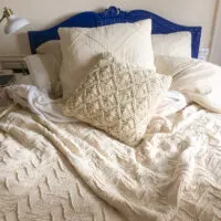 A bed with an ornate blue headboard is made up with a cream colored duvet cover and textured pillows.