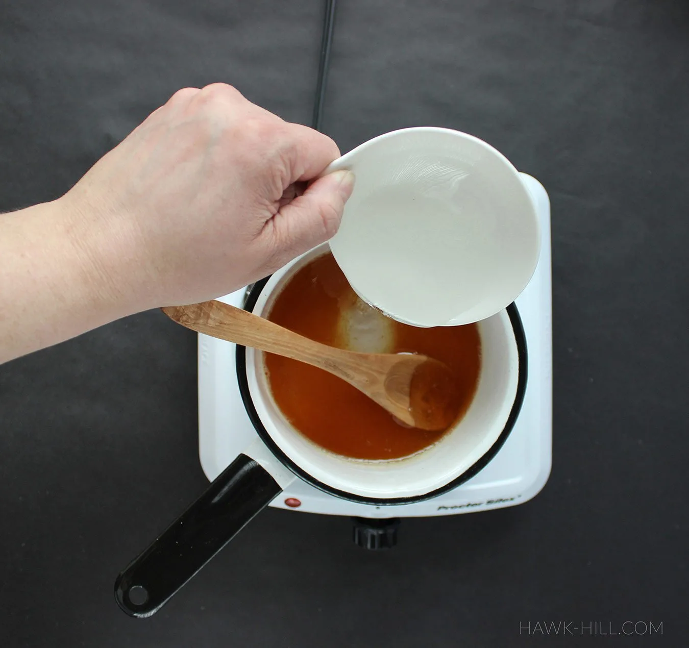 A hand pours a small amount of corn syrup into a pan of honey in order to prevent crystalization.