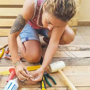 A young adult woman with a tattoo is shown kneeling next to tools and holding a project in her hands.