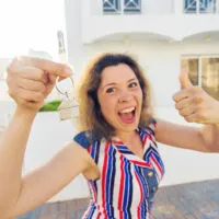 A woman in a striped shirt poses in front of a house with a thumbs up while holding keys in her opposite hand.