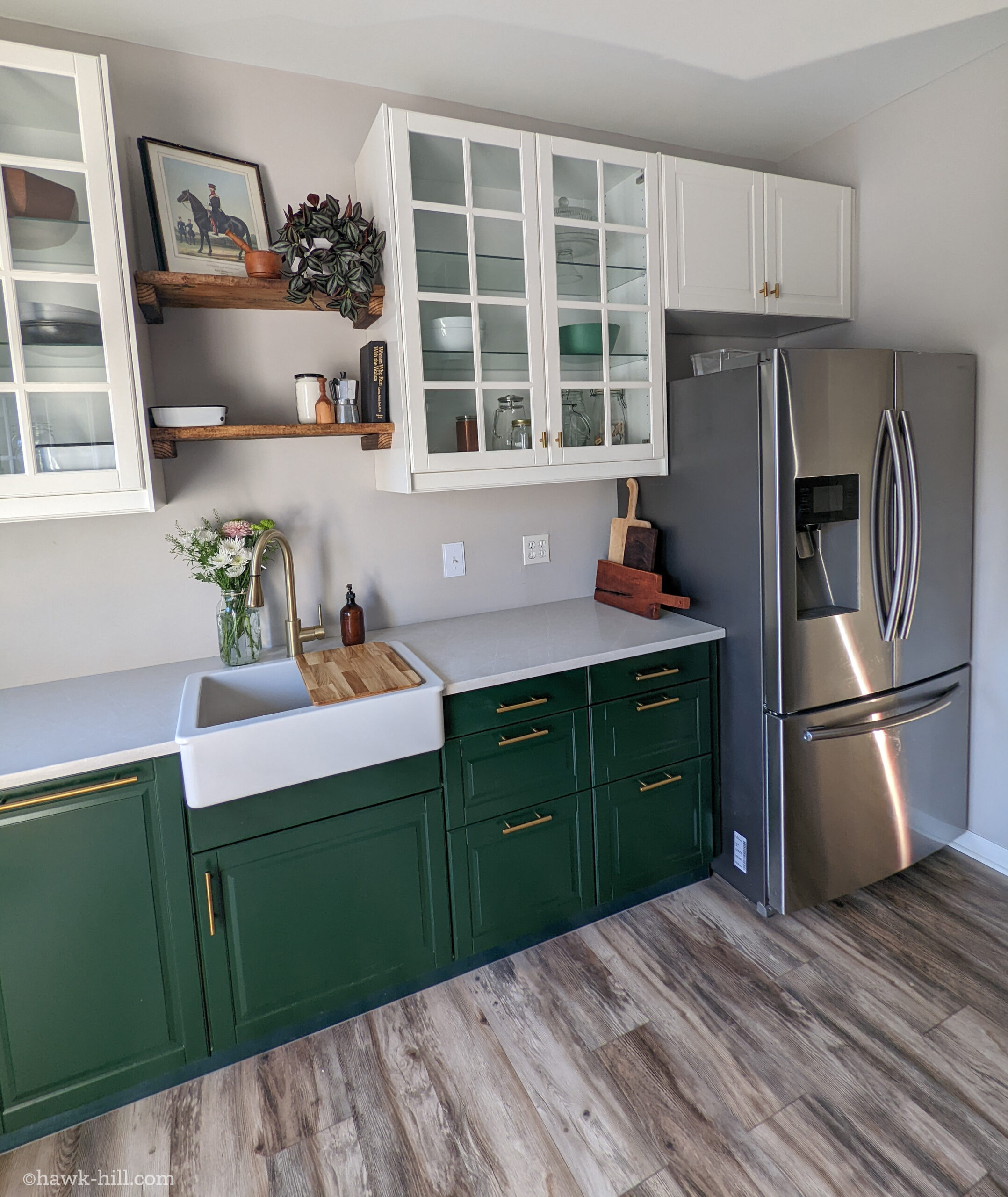 A kitchen with gold fixtures, forest green cabinets on the bottom, and a combination of wood open shelving and glass front white cabinets on top.
