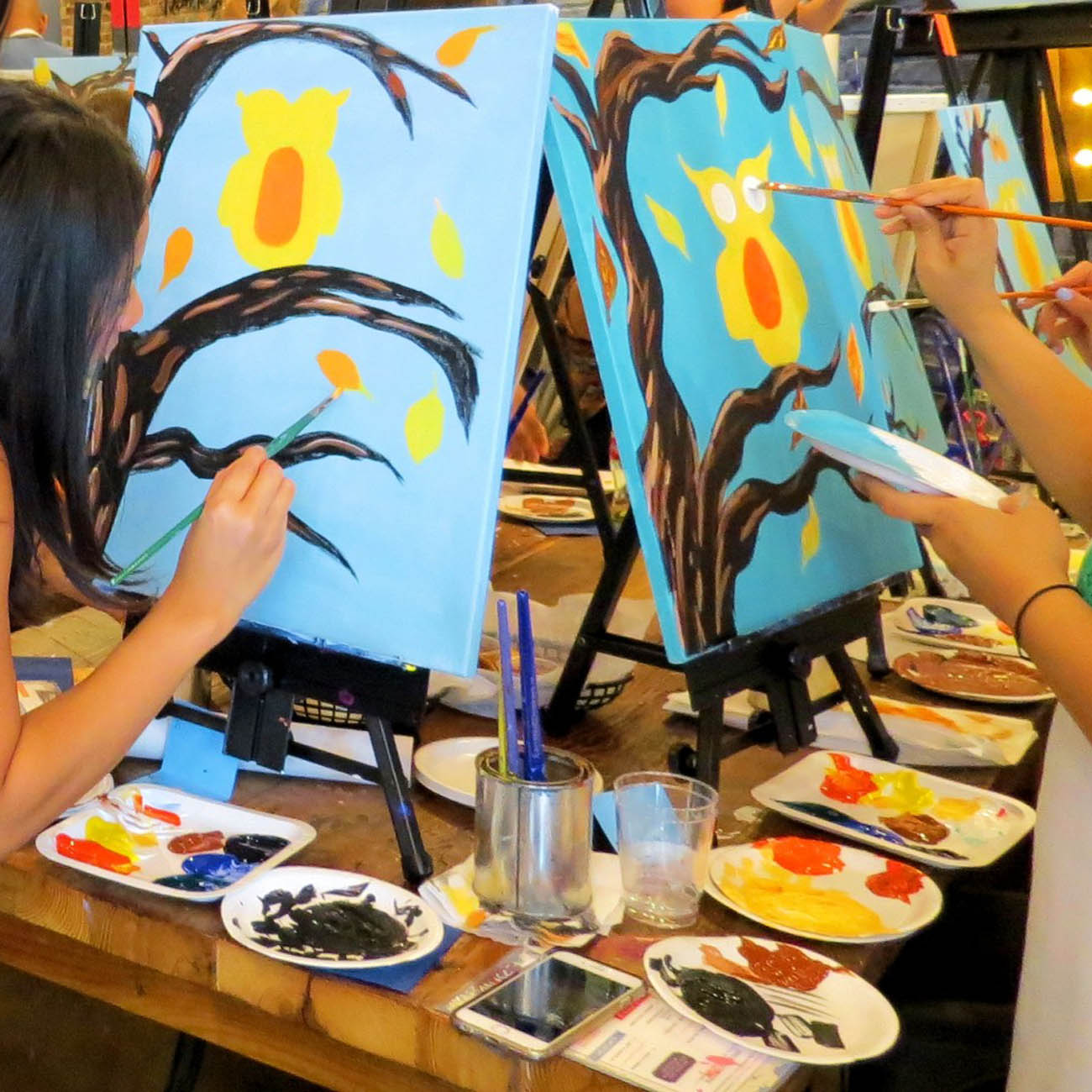 partygoers paint owls on blue canvases at a paint and sip party.